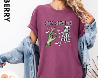 以下が含まれることがあります： ベリー色の半袖Tシャツ。緑色の手と白い骸骨の手がOKサインをしているグラフィックがプリントされています。その上には「SIX SEVEN」という文字があり、小さな白いコウモリと星型のきらめきが周囲に描かれています。