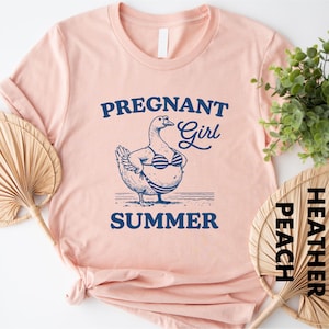 a pink t-shirt with a graphic design of a duck wearing a bikini top and the text "PREGnant Girl SUMMER" printed on it. The t-shirt is placed on a white surface, with a potted plant and a fan visible in the background.
