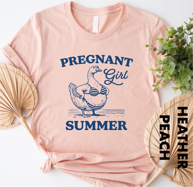 a pink t-shirt with a graphic design of a duck wearing a bikini top and the text "PREGnant Girl SUMMER" printed on it. The t-shirt is placed on a white surface, with a potted plant and a fan visible in the background.