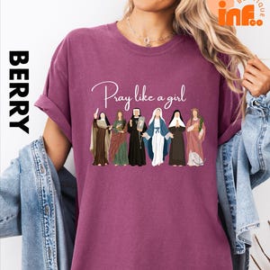 A woman wearing a purple t-shirt with a graphic design of various religious figures, including saints and religious icons, along with the text "Pray like a girl".