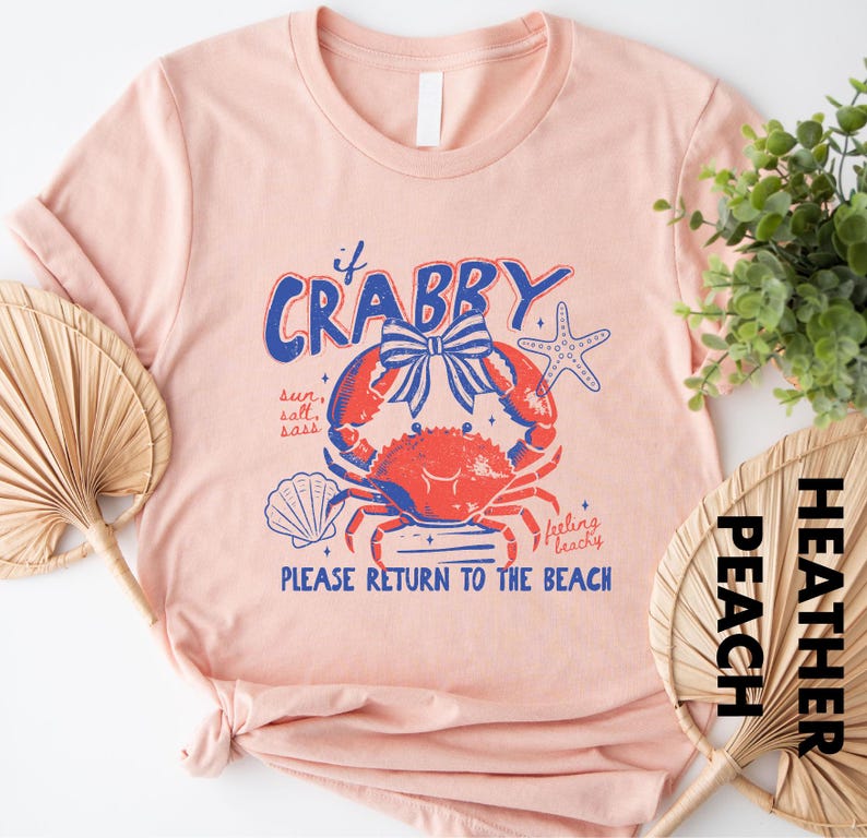 a pink t-shirt with a crab design and text, along with a potted plant and a fan, suggesting a beach or coastal theme.