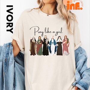 A woman wearing a white t-shirt with a graphic design of six different women in various religious attire, along with the text "Pray like a girl".