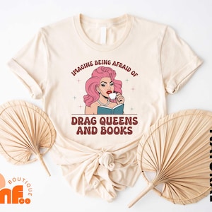 May include: Cream-colored t-shirt featuring a graphic of a drag queen with pink hair, drinking from a cup while reading a book. The text reads "IMAGINE BEING AFRAID OF DRAG QUEENS AND BOOKS."