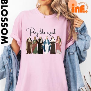 A woman wearing a pink t-shirt with a graphic design of six women in religious attire, along with the text "Pray like a girl".