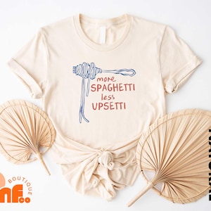 May include: A beige t-shirt with a blue and red graphic of a fork and spaghetti. The text on the shirt reads "more spaghetti less upsetti".