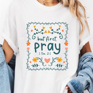 May include: White t-shirt with a floral design and the text "but first, pray" in green. The design includes orange flowers and a heart. The text "1 Tim. 2:1" is below the word "pray".