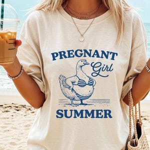 A woman wearing a white t-shirt with a graphic design of a duck wearing a bikini top and the text "Pregnant Girl Summer" printed on it, standing on a beach with a straw drink in her hand.