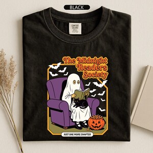 May include: A black t-shirt featuring a graphic of a ghost in a white sheet sitting in a purple armchair reading a book with a black cat on its lap. The design includes bats, a jack-o'-lantern, and the text The Midnight Readers Society and JUST ONE MORE CHAPTER.