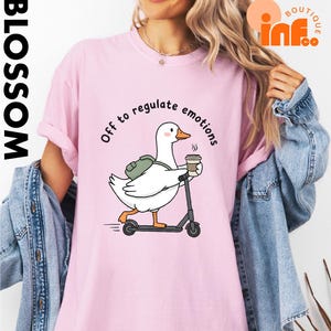 May include: Pink t-shirt with a cartoon goose riding a scooter and holding a coffee cup. The goose has a backpack and the text "Off to regulate emotions" is above the goose. The word "BLOSSOM" is printed vertically on the left side.