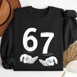 May include: Black sweatshirt with the number 67 in white, along with two open hands in a line drawing style. The word "BLACK" is printed vertically on the right side of the sweatshirt.