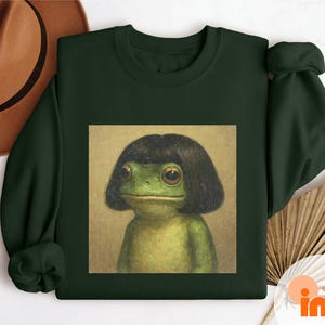 May include: Forest green sweatshirt featuring a frog with a black wig. The frog is depicted in a square image on the front of the sweatshirt. The text "FOREST GREEN" is printed vertically on the right side of the sweatshirt.