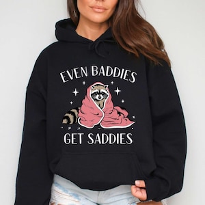 May include: Black hooded sweatshirt with the text "Even Baddies Get Saddies" and a cartoon raccoon wearing a pink blanket.