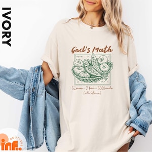 May include: Ivory-colored t-shirt with the text "God's Math" in brown, featuring a green illustration of fish and bread. The text "5 Leaves - 2 fish = 5000 meals (with leftovers)" is below the illustration. The shirt is worn with a blue denim jacket.