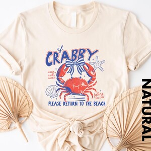 A beige t-shirt with a crab design and text, along with two palm fronds, suggesting a beach or coastal theme.
