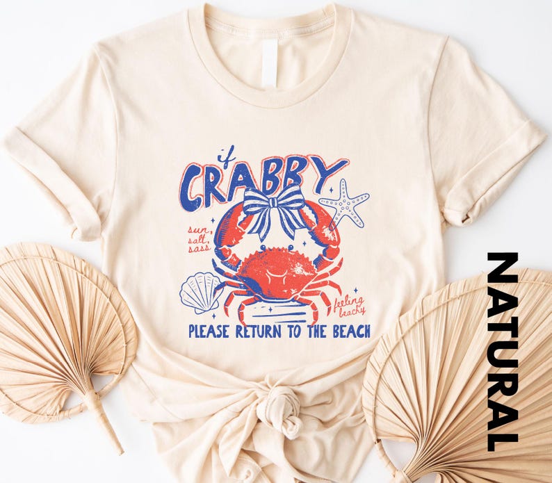 A beige t-shirt with a crab design and text, along with two palm fronds, suggesting a beach or coastal theme.