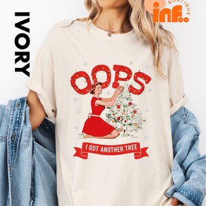 May include: Ivory-colored t-shirt featuring a retro illustration of a woman in a red dress decorating a Christmas tree. The word "OOPS" is above the illustration, and the text "I GOT ANOTHER TREE" is on a banner below.