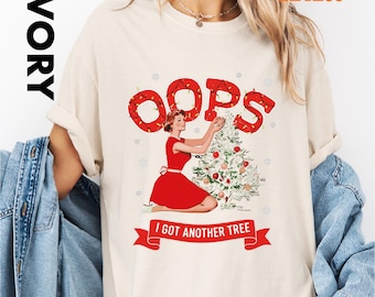 May include: Ivory-colored t-shirt featuring a retro illustration of a woman in a red dress decorating a Christmas tree. The word "OOPS" is above the illustration, and the text "I GOT ANOTHER TREE" is on a banner below.