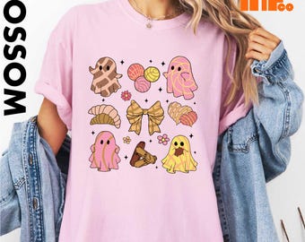 May include: A light pink short-sleeve t-shirt featuring a graphic print of three ghosts, various Mexican sweet breads like conchas, a bow, and small flowers. The design includes brown, pink, and yellow colors with small star accents throughout.