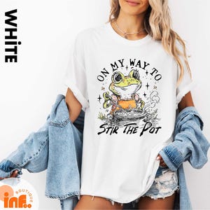 May include: White t-shirt with a graphic of a frog stirring a pot, with the text "ON MY WAY TO STIR THE POT". The frog is wearing an orange apron. The shirt has rolled-up sleeves.