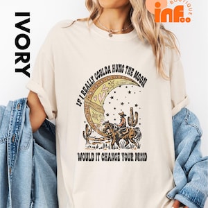 May include: Ivory-colored t-shirt featuring a graphic of a cowboy riding a horse in front of a crescent moon, stars, and cacti. The text reads "IF I REALLY COULDA HUNG THE MOON WOULD IT CHANGE YOUR MIND".