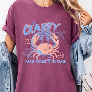 A woman wearing a purple t-shirt with a crab design and text, standing in front of a denim jacket.