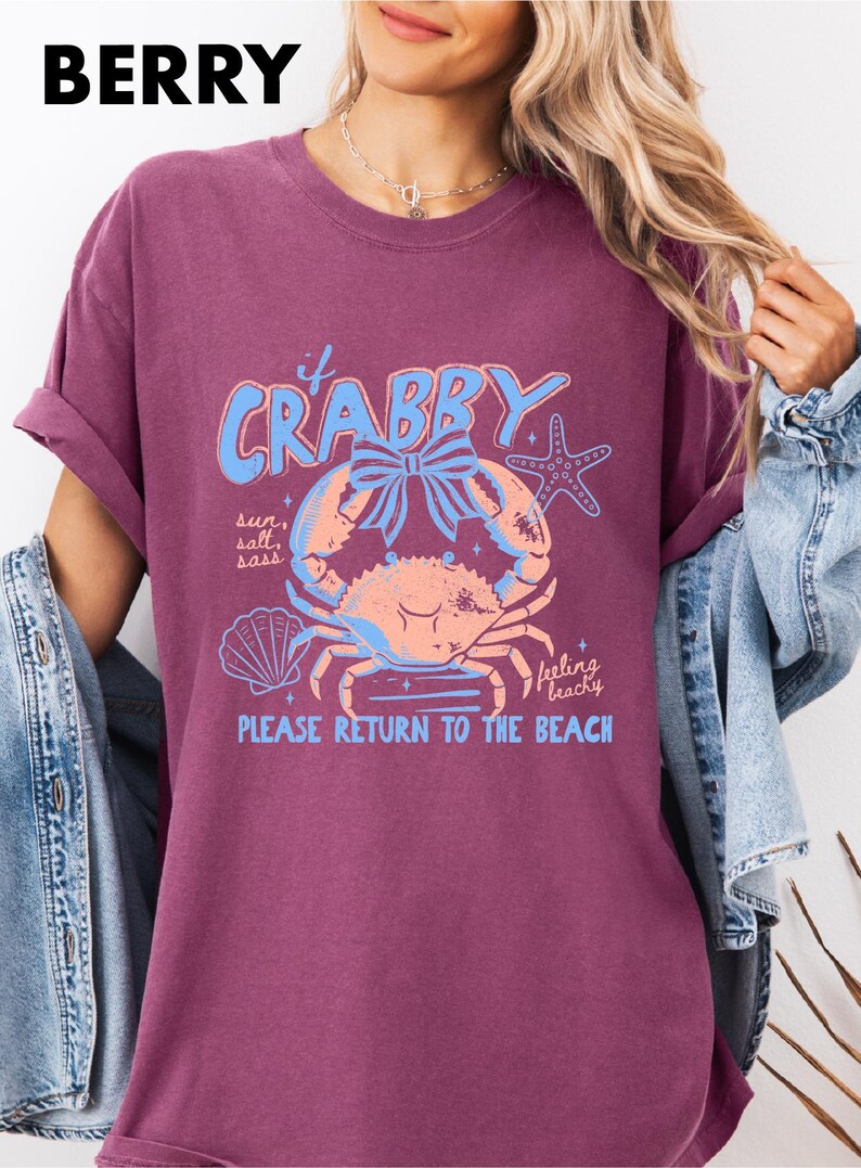 A woman wearing a purple t-shirt with a crab design and text, standing in front of a denim jacket.