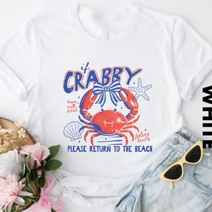 a white t-shirt with a crab design and text, along with a pair of sunglasses, a bouquet of flowers, and a pair of jeans, all arranged on a white background.