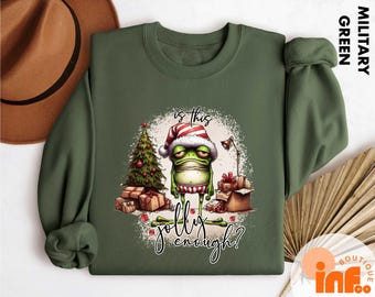 May include: Military green sweatshirt featuring a grumpy frog wearing a Santa hat, surrounded by Christmas gifts and a tree. The text "Is this jolly enough?" is written in a script font.