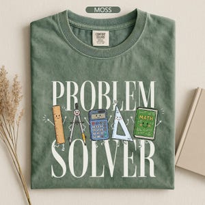May include: A moss green t-shirt featuring the white text "PROBLEM SOLVER" and a graphic of five cartoon school supplies including a ruler, compass, calculator, triangle, and a green math book, all with smiling faces and stick-figure arms and legs.