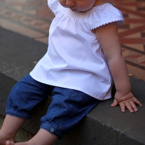 May include: A young child wearing a white, ruffled blouse with short sleeves and blue denim shorts. The child is sitting on a concrete step and looking down.