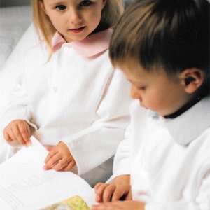 May include: Two children in white pajamas are sitting on a bed, reading a book together. The girl is wearing a pink collar on her pajamas.