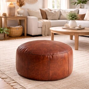 May include: A round, brown leather ottoman sits on a beige rug in a living room setting. The ottoman has a stitched design and is the focal point of the room. The rug has a fringed edge, and a white sofa and wooden furniture are in the background.