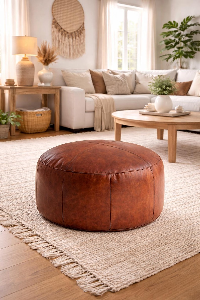 May include: A round, brown leather ottoman sits on a beige rug in a living room setting. The ottoman has a stitched design and is the focal point of the room. The rug has a fringed edge, and a white sofa and wooden furniture are in the background.