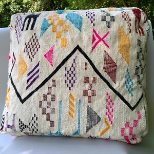 May include: A square throw pillow featuring an off-white woven fabric base decorated with embroidered geometric patterns in vibrant colors including hot pink, bright blue, yellow, purple, and black, centered by a prominent black zigzag line.