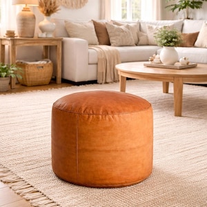 May include: A round, brown leather ottoman sits on a beige rug in a living room setting. The ottoman is a warm, caramel color and has a smooth, slightly textured surface. The room features a white sofa, wooden tables, and natural light.