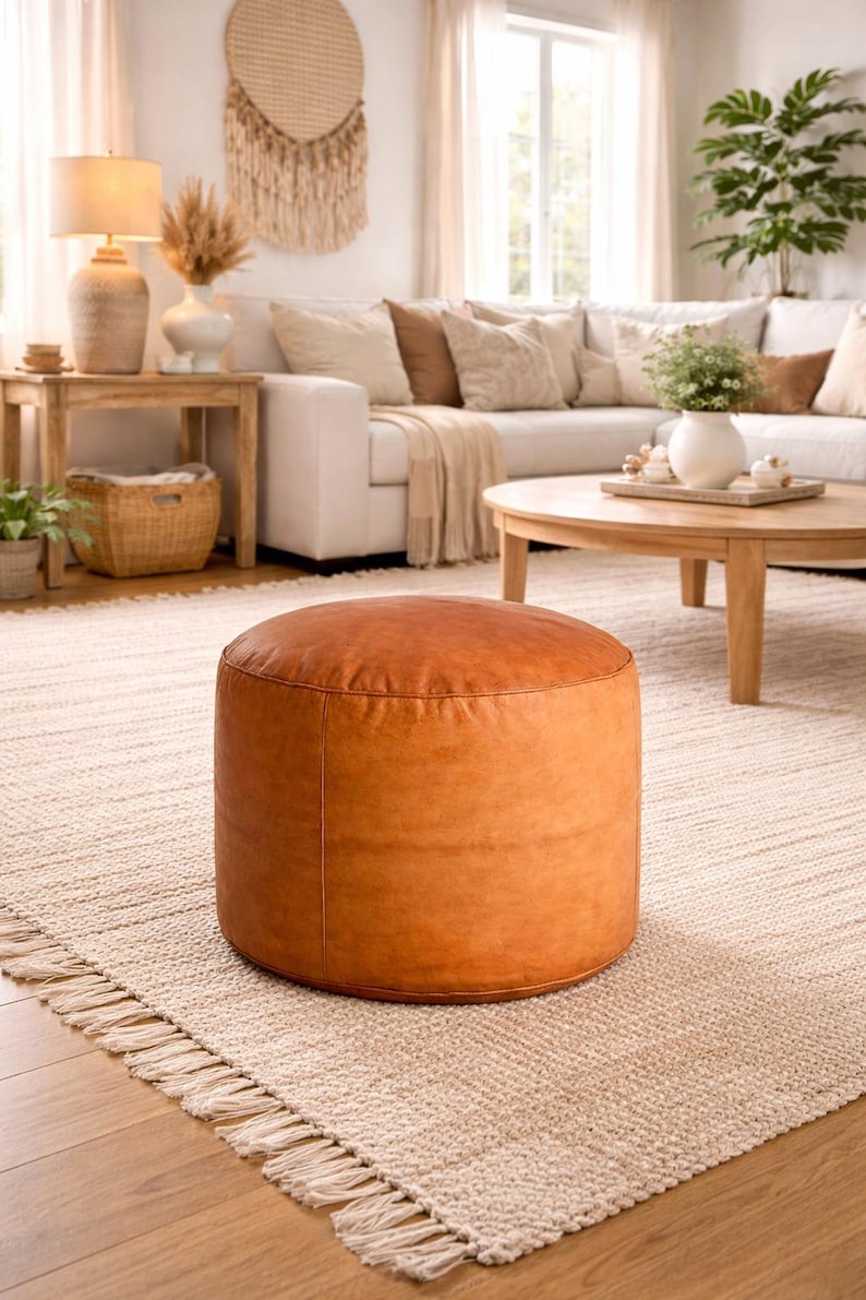 May include: A round, brown leather ottoman sits on a beige rug in a living room setting. The ottoman is a warm, caramel color and has a smooth, slightly textured surface. The room features a white sofa, wooden tables, and natural light.