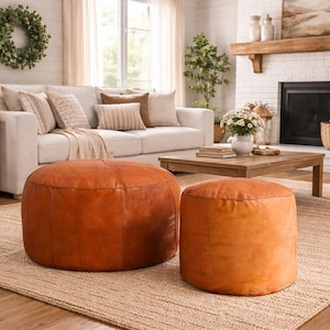 May include: Two round, brown leather ottomans of different sizes sit on a woven rug in a living room setting. A white sofa with decorative pillows and a wooden coffee table are in the background. The room has a neutral color palette.