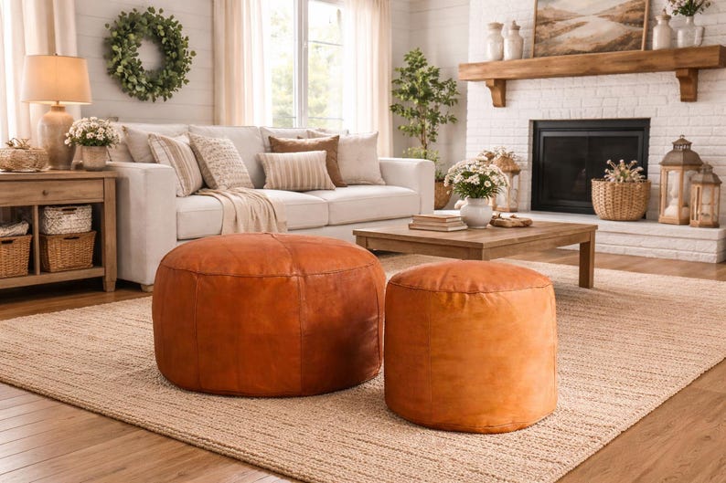 May include: Two round, brown leather ottomans of different sizes sit on a woven rug in a living room setting. A white sofa with decorative pillows and a wooden coffee table are in the background. The room has a neutral color palette.