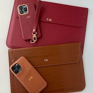 May include: Two personalized leather accessories: a burgundy laptop sleeve and phone case with gold initials "T.M" and "M.F". A brown, crocodile-textured laptop sleeve and phone case with initials "J.M" and "A.W" are also shown.