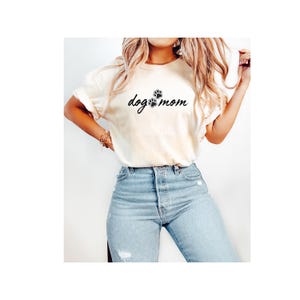 Puede incluir: Camiseta color crema con la frase "dog mom" en fuente manuscrita, acompañada de una huella de pata. La camiseta se combina con vaqueros azul claro.