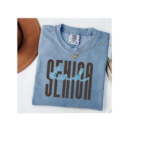 Puede incluir: Una camiseta azul claro con las palabras "SENIOR dad" impresas en la parte delantera en marrón y verde azulado. La camiseta tiene cuello redondo y mangas cortas. Un collar dorado con un colgante está drapeado sobre la camiseta.