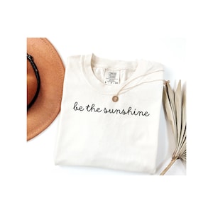 Puede incluir: Camiseta color crema con la frase "be the sunshine" en escritura negra. Un collar dorado con un colgante descansa sobre la camisa. Un sombrero marrón y hojas de palma secas también están en la imagen.
