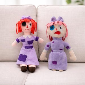 Op de afbeelding: Twee handgemaakte pluche poppen met gestikte kenmerken. De poppen hebben rood garen haar, paarse jurken met patches en ongelijke ogen. Eén pop heeft een zwart oog en de andere een blauw oog. Ze zitten op een lichtgrijze bank.