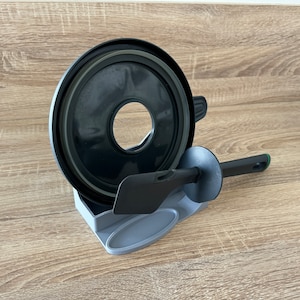 Pode incluir: Uma tampa preta de aparelho de cozinha com uma abertura central, uma espátula preta e um suporte cinza. A espátula tem um cabo preto com um detalhe verde. Os itens estão em uma superfície de madeira.