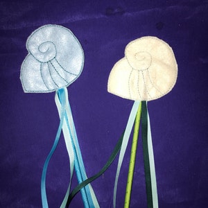May include: Two seashell wands with ribbon streamers. One is light blue, the other off-white. Both have embroidered details and are set against a dark purple background. Ribbons are shades of blue, green, and black.