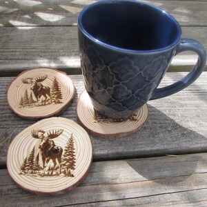 May include: Three wooden coasters with a burnt-in design of a moose standing in a forest. The coasters are round and have a natural wood finish. A blue ceramic mug is sitting on one of the coasters.