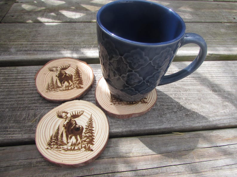 May include: Three wooden coasters with a burnt-in design of a moose standing in a forest. The coasters are round and have a natural wood finish. A blue ceramic mug is sitting on one of the coasters.