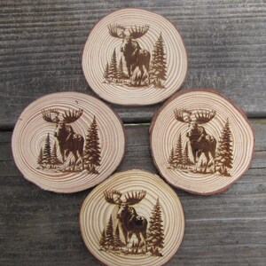 May include: Set of four wooden coasters featuring a moose design. Each coaster is circular and has a natural wood finish with a dark brown moose silhouette burned into the surface.