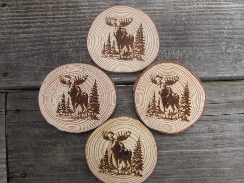 May include: Set of four wooden coasters featuring a moose design. Each coaster is circular and has a natural wood finish with a dark brown moose silhouette burned into the surface.