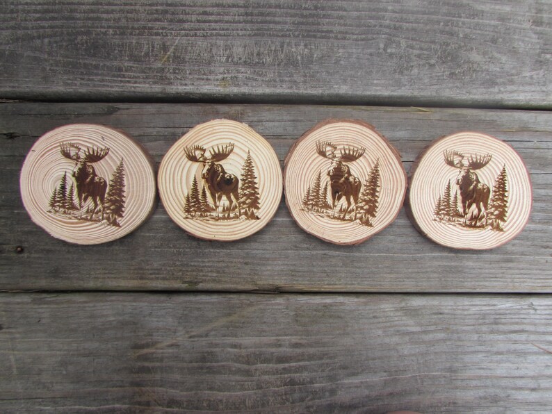 May include: Four wooden coasters with a burnt design of a moose standing in a forest. The coasters are round and have a natural wood finish.